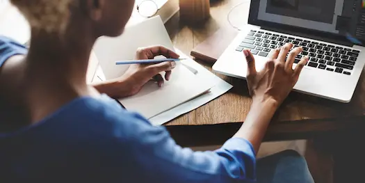 Woman sitting at a desk working on a laptop and taking notes with pen and paper