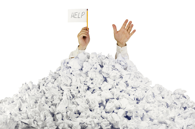 Image of person buried under a pile of papers holding a help sign