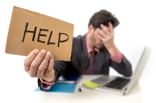 young businessman in suit and tie sitting at office desk working on computer laptop asking for help holding cardboard sign looking sad and depressed