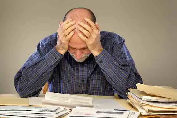 A senior adult man sitting at a table or desk stacked with papers and envelopes looking down with his hands on his head