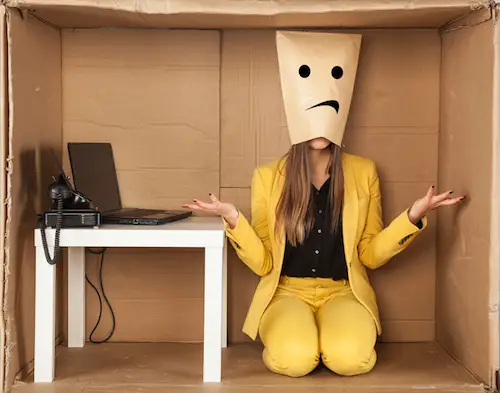 Sad business woman donned a paper mask sitting inside of a box next to a workbench with a laptop