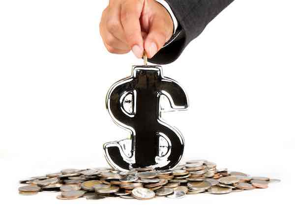 Businessman's hand depositing a coin into a dollar sign shaped piggy bank surrounded by a pile of coins