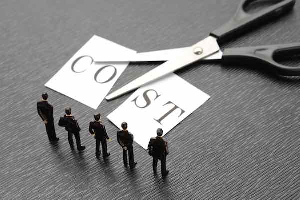 Word 'cost' cut in half with scissors lying next to it as a group of businessmen stand looking at it