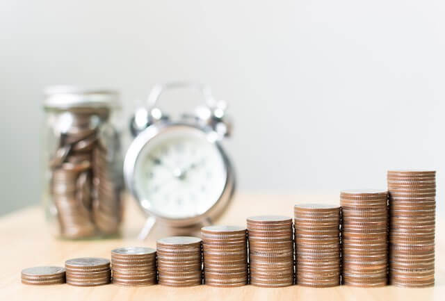 Stacks of coins getting progressively larger in size in the foreground with a blurred image of an old style alarm clock and jar full of money in the background depicting saving for retirement and the future