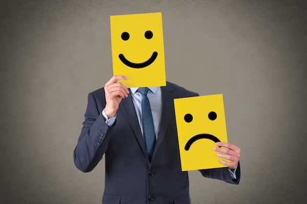 Businessman holding a happy face drawn on yellow cardboard in front of his face while holding a frowning face on yellow cardboard in his other hand held at a lower height