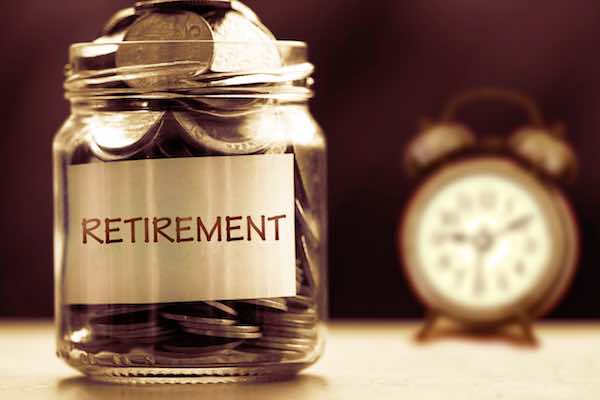 Close up of coins in a glass jar labeled 'retirement' with an alarm clock in the background