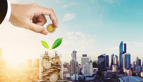 Close up of a businessman's hand putting coins in a jar overlaid against a city skyline depicting a locality pay concept