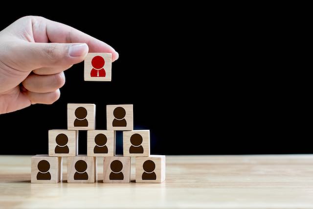 Closeup of a person's hand stacking wooden blocks each containing an icon depicting a person into a pyramid shape depicting performance and human resources management