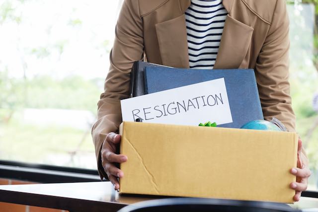 Businesswoman carrying a box of her personal belongings out of an office with a form clearly labeled 'resignation' showing in the forefront of the box contents