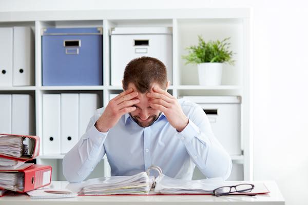 Young businessman sitting at his desk appearing stressed out holding his hands to his forehead as he looks downward at a binder of paperwork