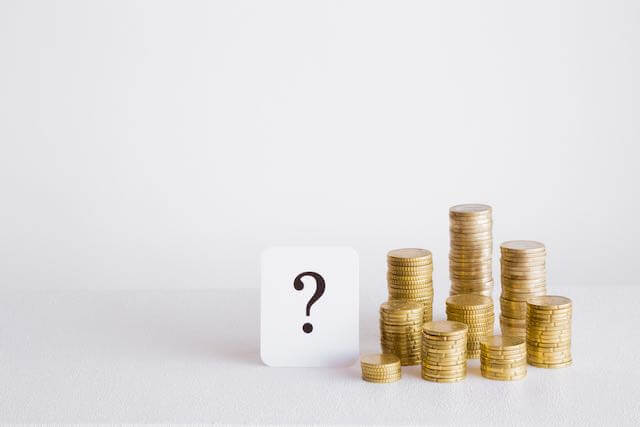 Black question mark on a white index card sitting upright next to a collection of small stacks of coins depicting confusion about money or salary