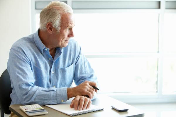 Senior citizen aged man sitting at a desk with a pen, notepad and calculator staring off into space as he appears to think deeply about the future