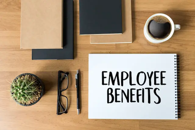 White sheet of copy paper with words 'employee benefits' printed on it sitting on a wooden desk next to a pen, glasses, coffee cup and books