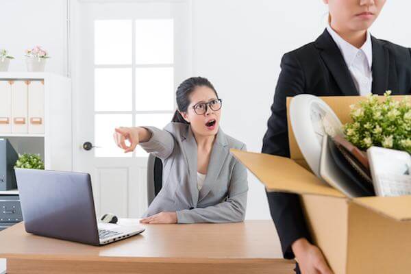 Woman sitting at her desk pointing towards the door appearing to tell an employee carrying a box of her personal belongings that she has been fired