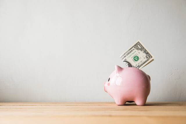 Piggy bank on a wood table with a dollar bill folded and sticking out of the coin slot in the piggy bank's back