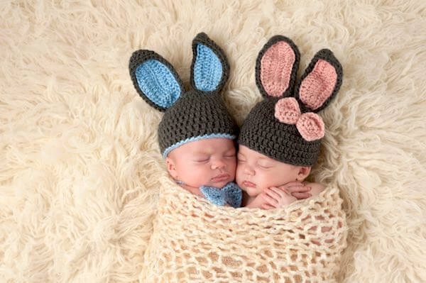 Newborn boy and girl wearing blue and pink rabbit ear hats lying asleep wrapped in a wool blanket