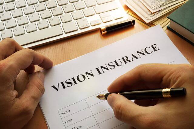 Close up of a person's hands filling out a form labeled 'vision insurance' sitting at a desk with a computer keyboard in the background