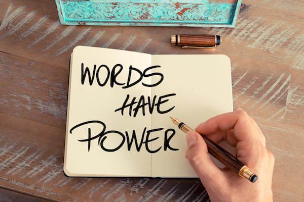 Close up of a person's hand writing 'words have power' in a book sitting on a wooden desk