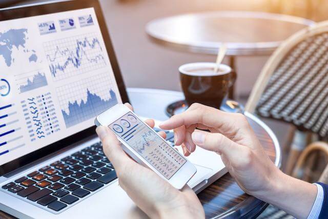 Close up of a person's hands holding a smartphone to check stock market performance graphs with a laptop in the background also displaying financial charts