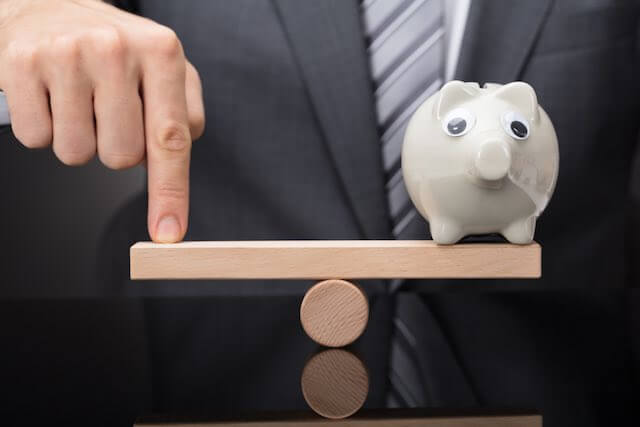 Businessman's finger on one end of a seesaw with a piggy bank on the other to balance it out and make it level; pay parity concept