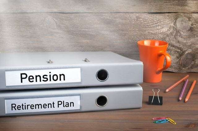 Two binders stacked on top of each other on a desk next to a paper clip and coffee cup, one labeled 'pension', the other labeled 'retirement plan'
