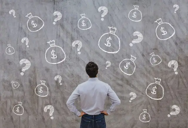 Man staring at a board with multiple drawings of bags of money and question marks depicting questions or confusion about money/personal finance/retirement