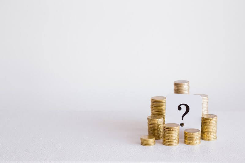 Stacks of gold coins around a question mark on a white index card all set against a white background