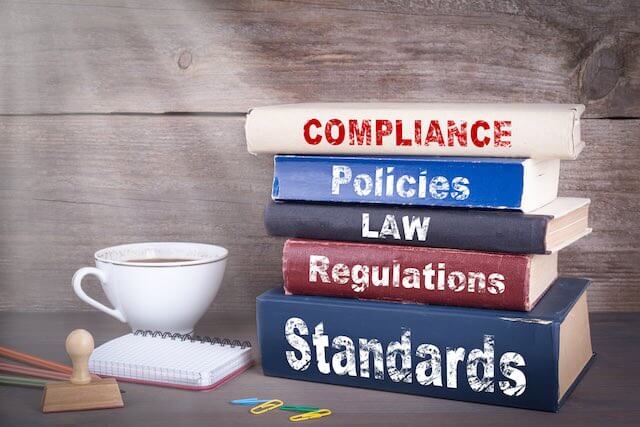 Stack of five books on a wooden desk next to a coffee cup in an office setting labeled 'compliance', 'policies', 'law', 'regulations', 'standards'
