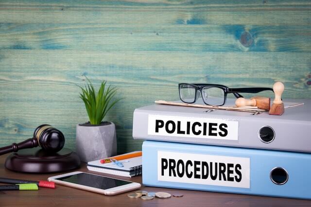 Two notebooks on a desk workspace labeled 'policies' and 'procedures' with a smartphone, pens and glasses