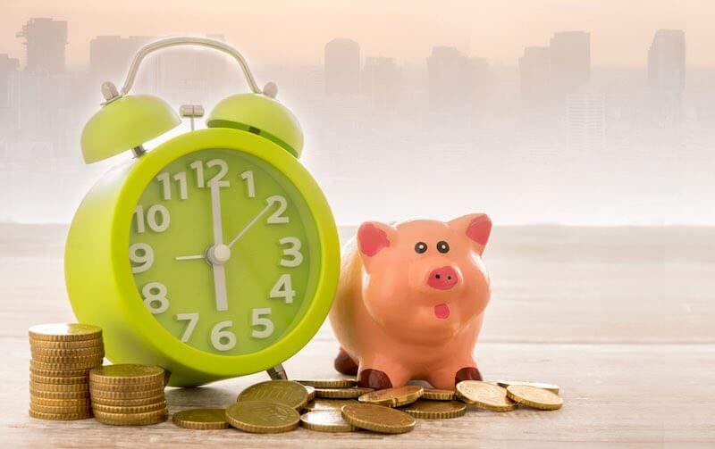 Old style alarm clock on a table next to a scattering of coins and a pink piggy bank depicting growing wealth over time/savings for retirement