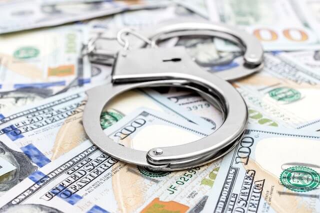 A pair of handcuffs sits on top of a spread of $100 cash bills depicting fraud/stealing/theft of public funds