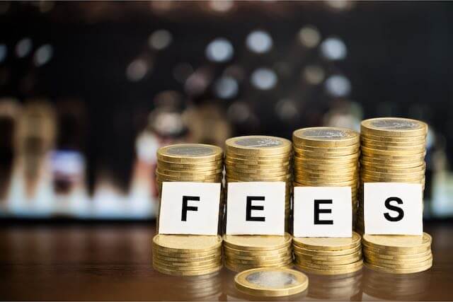 Vertical stacks of gold coins with the letters 'fees' on small white cardboard squares spelled out in front of the coin stacks depicting investment fees or transaction costs