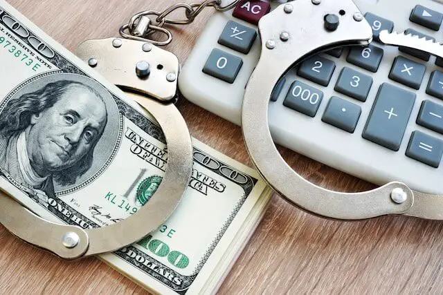 Stack of $100 bills on a desk next to a calculator and handcuffs depicting identity theft/account/financial fraud