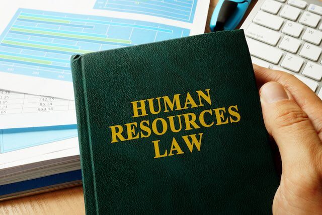 close up of person's hand holding a book titled 'human resources law' with desk and computer keyboard in background