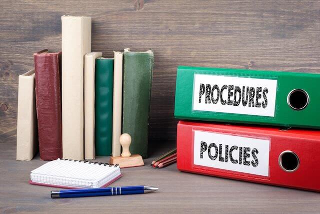 Two binders on a desk against a wooden backdrop labeled 'policies' and 'procedures' next to a notepad and pen depicting human resources/workplace disciplinary procedures
