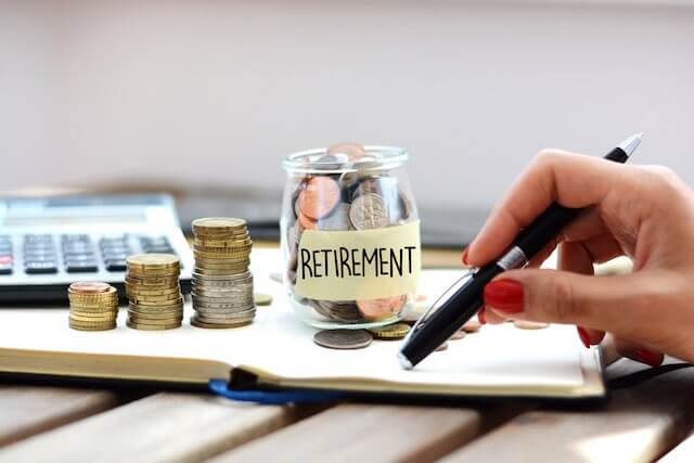 Vertical stacks of coins increasing in size leading up to a jar full of coins labeled 'retirement' pictured beside a close up of a woman's hand holding a pen pondering her future retirement savings/taxes/nest egg