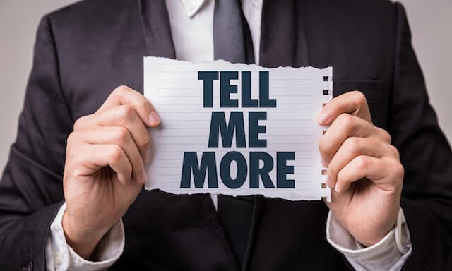 Businessman holding a torn sheet of paper up that reads 'tell me more' depicting public speaking skills/credibility/listening