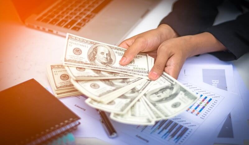 Close up of a woman's cash holding a spread of $100 cash bills over a desk with budget forms, a laptop and a notebook