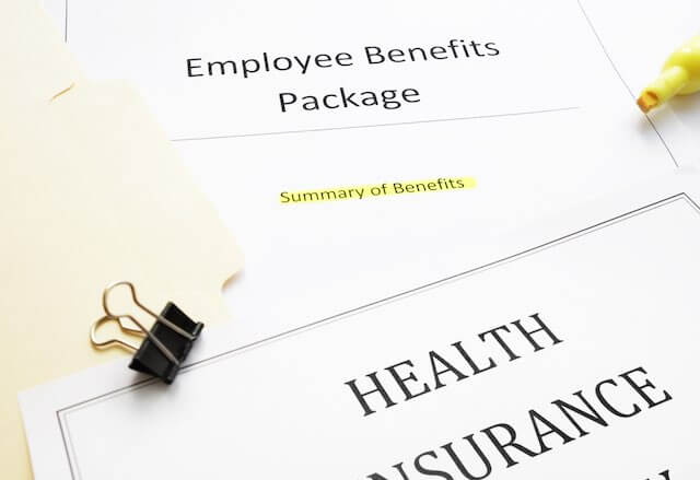 Paperwork on a desk labeled 'employee benefits package' with a document in the foreground that reads 'health insurance'