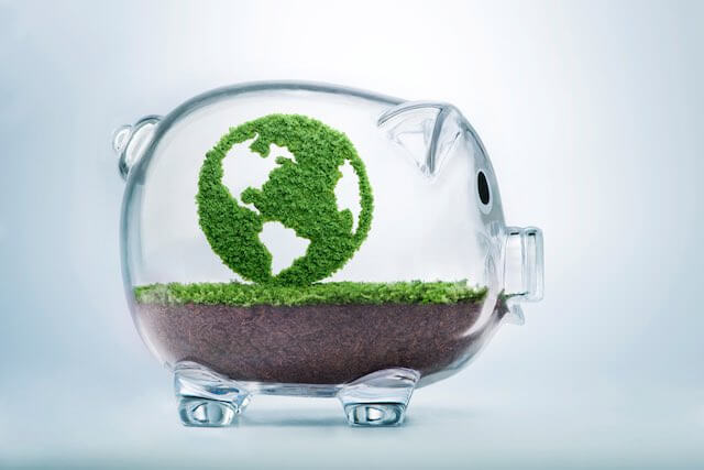 Clear glass piggy bank filled with a layer of dirt and grass with a grass symbol of the earth inside of it depicting green or socially responsible spending/investing