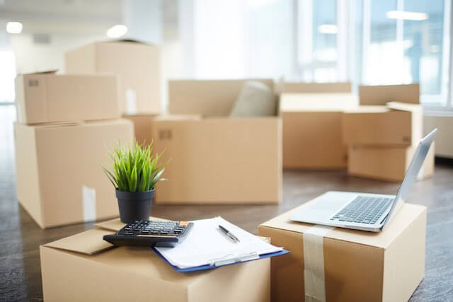 Stacks of cardboard moving boxes with a laptop and clipboard sitting on top of some of them depicting moving or a job relocation or job transfer