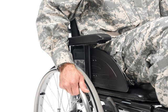 Close up of the side of a wheelchair with a male veteran sitting in it wearing a camouflage uniform with his hand gripping the wheel of the wheelchair - disability, disabled veteran, va benefits