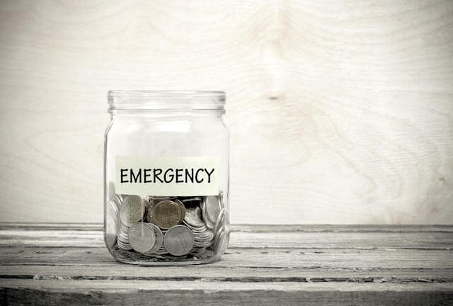 Jar of coins labeled 'emergency' sitting on a wooden table depicting an emergency fund/emergency savings