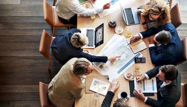 Group of employees seen from above sitting around a conference room table collaborating on a project as they work on laptops and review charts and paperwork