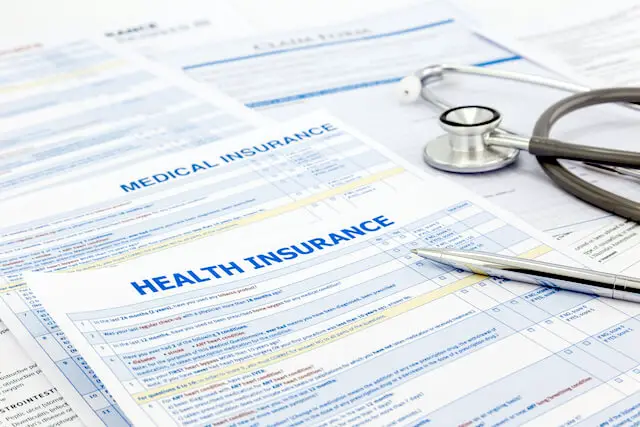 Series of documents labeled 'health insurance' and 'medical insurance' with a pen and stethoscope lying on top of them; FEHB plans and paperwork