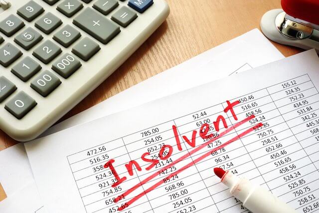 Word 'insolvent' written in large red letters across a spreadsheet on a desk next to a calculator depicting bankruptcy or dire finances