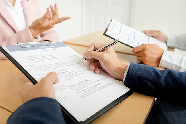 Close up of the files and clipboards held by two managers sitting on one side of a conference room table interviewing a businesswoman job candidate on the opposite side