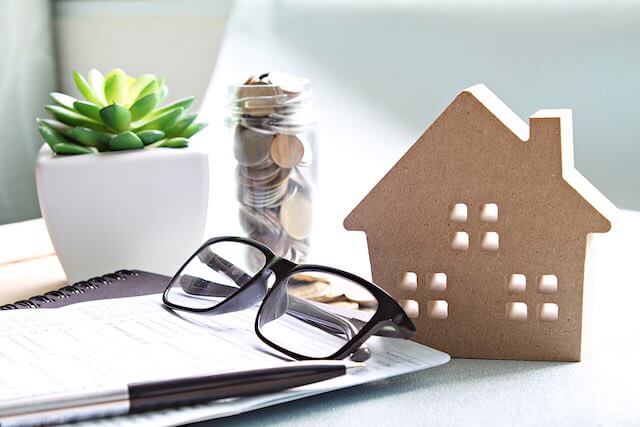 Small wooden carving of a house on a desk next to a plant, jar of coins, notebook, pen and glasses depicting home mortgage debt/financial planning
