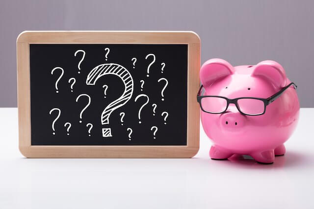 Pink piggy bank wearing glasses next to a chalkboard with multiple question marks