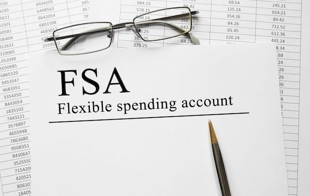 Piece of paper labeled 'flexible spending account' lying on top of a spreadsheet next to a pen and reading glasses depicting planning for health care/health insurance expenses